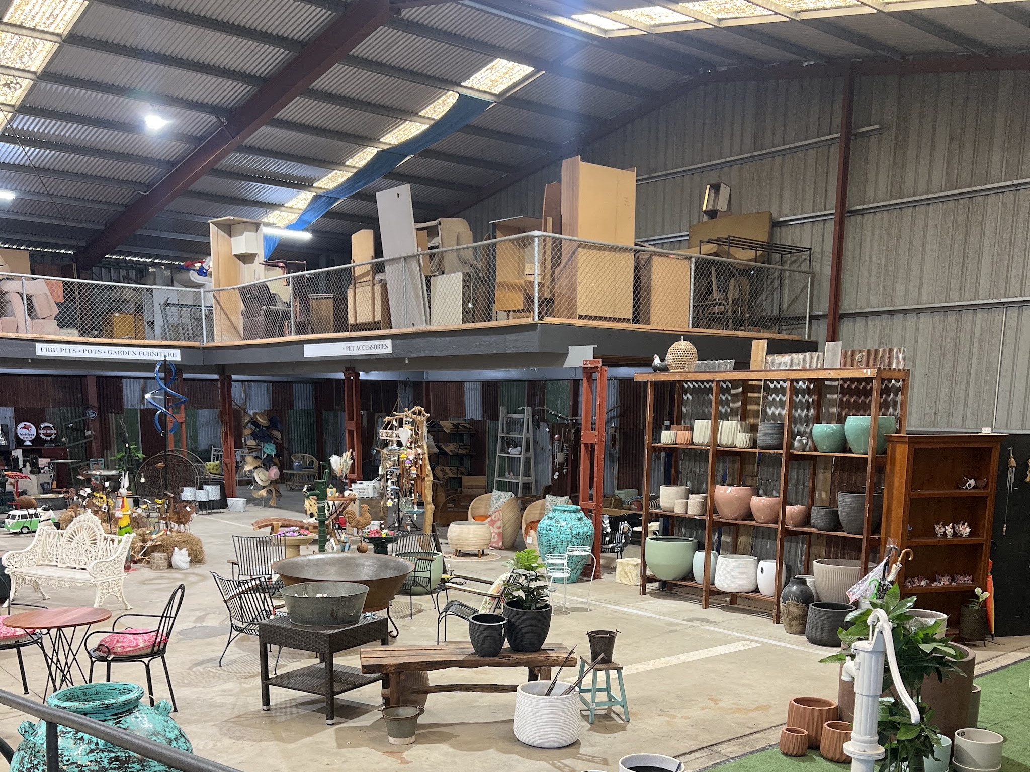 Wide view across the covered market shed, showing the mezzanine, the fire pits and garden furniture signage, the pot wall and the furniture floor